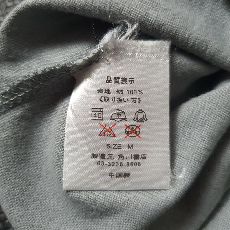 May include: White tag with black text and symbols. The tag says "Quality Display", "Surface Cotton 100%", "<Handling Method>", "40", "Medium", "Size M", "Manufacturer:  Kadokawa", "03-3238-8606", "Made in China".