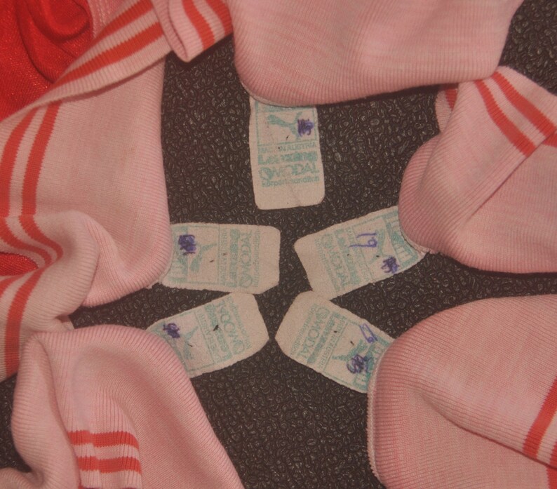 Op de afbeelding: Vier roze en rood gestreepte stofstalen met witte labels eraan bevestigd. De labels hebben blauwe en groene tekst die "SOCDA" en "LOS ANGELES, CALIFORNI&Euml;" luidt. E&eacute;n label heeft het nummer "19" erop geschreven.