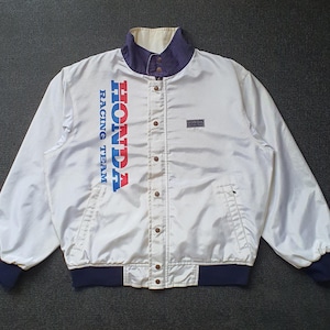 Peut inclure: Une veste blanche Honda Racing Team avec un col et des poignets bleu foncé. L'avant présente le texte HONDA RACING TEAM en lettres bleues et rouges, avec un petit écusson Honda sur la poitrine gauche et une fermeture à boutons pressions.