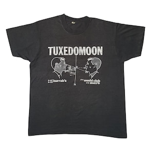 May include: Dark gray T-shirt with the band name "Tuxedomoon" in white block letters. The shirt features a graphic of two side profiles of men with lines and text. Below the graphic are the words "hurrah's" and "mudd club max's".