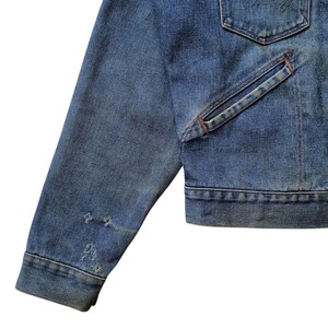 May include: Close-up of a blue denim jacket. The jacket has a classic design with a pocket and distressed details on the sleeve. The denim is a medium wash, showing signs of wear.