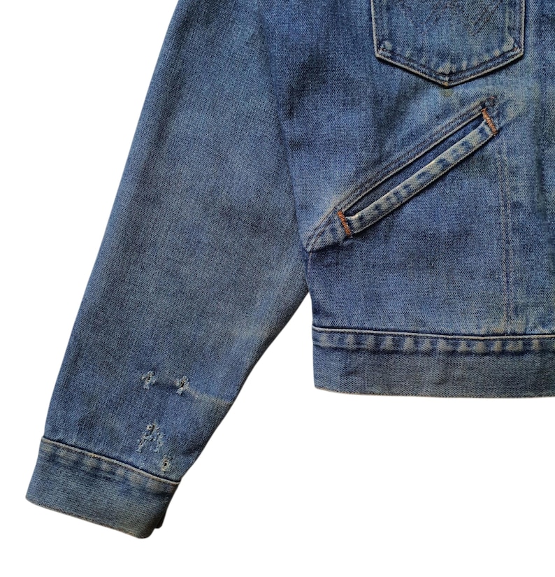 May include: Close-up of a blue denim jacket. The jacket has a classic design with a pocket and distressed details on the sleeve. The denim is a medium wash, showing signs of wear.