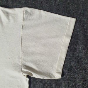 Pode incluir: Close-up do tecido de algod&atilde;o branco de uma manga de camiseta. O tecido &eacute; liso e tem uma textura sutil. A manga &eacute; costurada com uma costura reta.