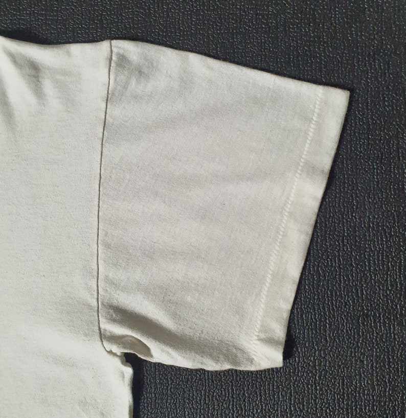 Pode incluir: Close-up do tecido de algod&atilde;o branco de uma manga de camiseta. O tecido &eacute; liso e tem uma textura sutil. A manga &eacute; costurada com uma costura reta.