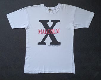 May include: White t-shirt featuring a large black "X" with the name "MALCOLM" in red across it. The shirt has a crew neck and short sleeves. Below the "X" is small text. The shirt is made of a soft material.