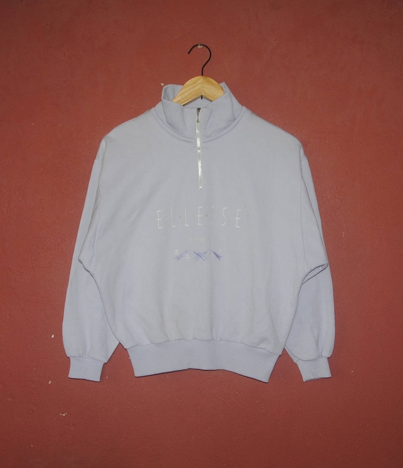May include: A light blue quarter-zip sweatshirt with the word "ellesse" embroidered on the front in white. The sweatshirt has a ribbed collar, cuffs, and hem.