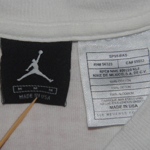 K&ouml;nnte beinhalten: Ein wei&szlig;es T-Shirt mit einem schwarz-wei&szlig;en Etikett, auf dem "Made in USA" steht und eine Silhouette eines Basketballspielers abgebildet ist. Das Etikett enth&auml;lt au&szlig;erdem die Texte "SP39-BAS", "RN# 56323", "CAR 05553", "RFC# NME 920220 K14", "NIKE DE MEXICO, SA DE CV", "100% BAUMWOLLE" und "MADE IN USA".