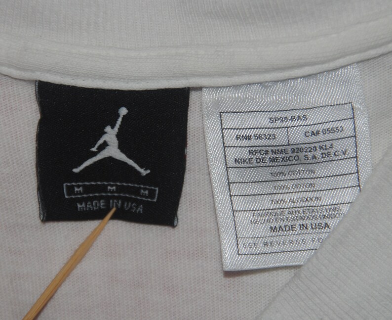 K&ouml;nnte beinhalten: Ein wei&szlig;es T-Shirt mit einem schwarz-wei&szlig;en Etikett, auf dem "Made in USA" steht und eine Silhouette eines Basketballspielers abgebildet ist. Das Etikett enth&auml;lt au&szlig;erdem die Texte "SP39-BAS", "RN# 56323", "CAR 05553", "RFC# NME 920220 K14", "NIKE DE MEXICO, SA DE CV", "100% BAUMWOLLE" und "MADE IN USA".