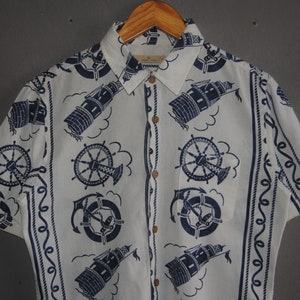 May include: A white short-sleeved button-down shirt with a nautical theme. The shirt features a repeating pattern of lighthouses, anchors, and ship wheels in navy blue on a white background. The shirt has a button-up collar and a button placket.