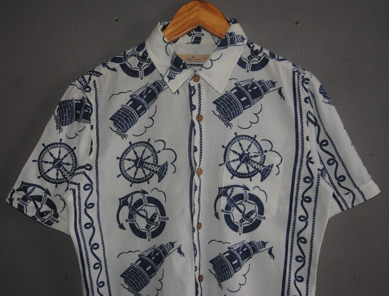 May include: A white short-sleeved button-down shirt with a nautical theme. The shirt features a repeating pattern of lighthouses, anchors, and ship wheels in navy blue on a white background. The shirt has a button-up collar and a button placket.