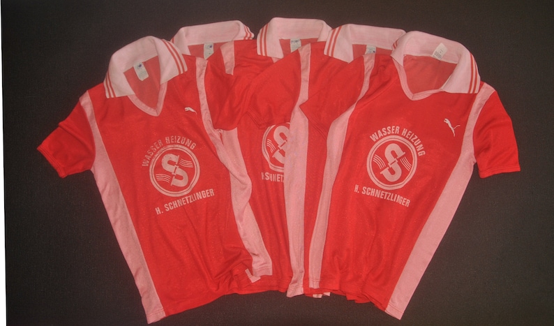 Op de afbeelding: Vijf rood-roze voetbalshirts met witte letters. De shirts hebben een witte kraag en witte strepen aan de zijkanten. De tekst op de shirts luidt "Wasser Heizung H. Schneitzlinger".