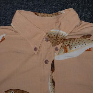 K&ouml;nnte beinhalten: Hellbraunes Button-Down-Hemd mit einem sich wiederholenden Muster von Fischillustrationen in Braun-, Wei&szlig;- und Oranget&ouml;nen. Der Kragen und die Kn&ouml;pfe sind sichtbar. Das Hemd ist aus leichtem Stoff gefertigt.
