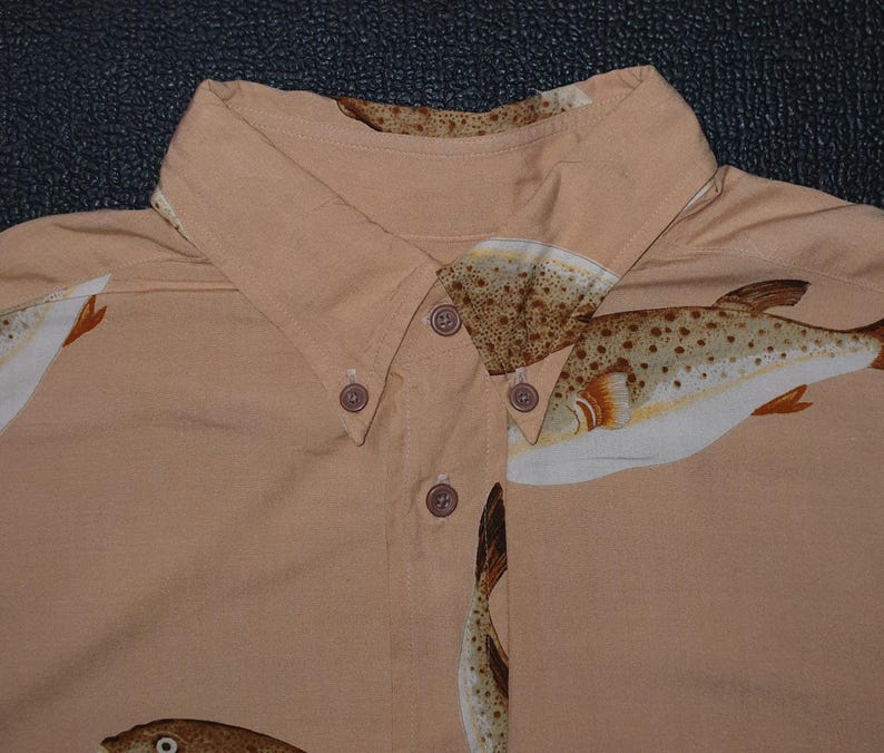 K&ouml;nnte beinhalten: Hellbraunes Button-Down-Hemd mit einem sich wiederholenden Muster von Fischillustrationen in Braun-, Wei&szlig;- und Oranget&ouml;nen. Der Kragen und die Kn&ouml;pfe sind sichtbar. Das Hemd ist aus leichtem Stoff gefertigt.
