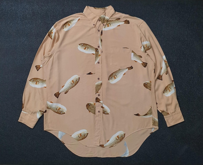 K&ouml;nnte beinhalten: Hellbeige Hemd mit Kn&ouml;pfen und einem sich wiederholenden Muster aus braunen und wei&szlig;en Kugelfischen. Das lang&auml;rmlige Hemd hat eine Brusttasche und einen klassischen Kragen.
