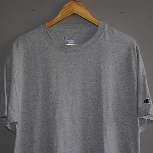 Op de afbeelding: Een grijs T-shirt met korte mouwen met een Champion-logo op de linkermouw en een Champion-label op de voorkant. Het label zegt "Champion" en "Reverse Weave" in witte tekst.
