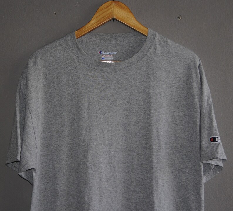 Op de afbeelding: Een grijs T-shirt met korte mouwen met een Champion-logo op de linkermouw en een Champion-label op de voorkant. Het label zegt "Champion" en "Reverse Weave" in witte tekst.