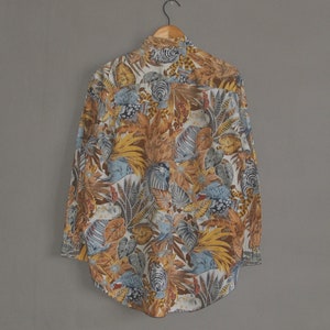 May include: A long-sleeved button-down shirt with a tropical print featuring a variety of leaves, flowers, and birds in shades of blue, brown, yellow, and white.