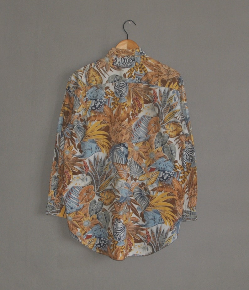 May include: A long-sleeved button-down shirt with a tropical print featuring a variety of leaves, flowers, and birds in shades of blue, brown, yellow, and white.