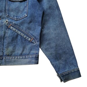 May include: Close-up of a vintage denim jacket in a medium wash. The jacket features a chest pocket, a slanted pocket, and a buttoned cuff. The denim shows signs of wear and fading, giving it a classic, worn look.