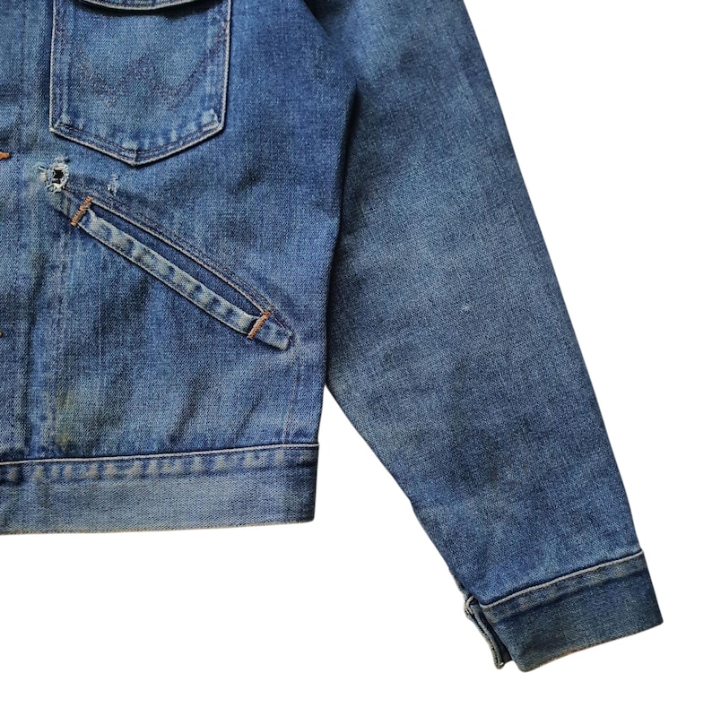 May include: Close-up of a vintage denim jacket in a medium wash. The jacket features a chest pocket, a slanted pocket, and a buttoned cuff. The denim shows signs of wear and fading, giving it a classic, worn look.