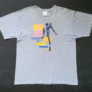 Könnte beinhalten: Graues T-Shirt mit einer blauen Silhouette eines Basketballspielers und dem Text "Nike Prep Classic San Diego" in Rosa, Gelb und Blau.