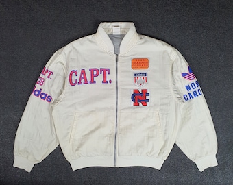 May include: A white vintage Adidas bomber jacket with a zipper closure. The jacket has several patches on the front, including a "CAPT." patch, a "1988 Champion Cougars" patch, an Adidas patch, and a North Carolina patch. The sleeves have the text "CAPT. 28 Adidas" on one sleeve and "North Carolina" on the other.