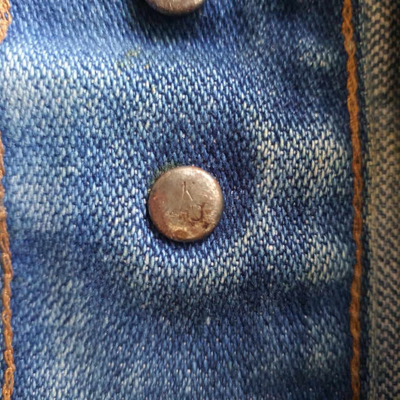 May include: Close-up of a section of blue denim fabric with two metal buttons. The buttons are round and have a slightly tarnished appearance. The denim has a classic, worn look with visible stitching.