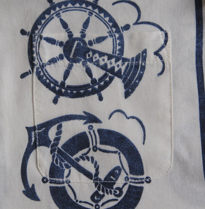 May include: A white fabric with a blue nautical design featuring a ship's wheel and an anchor. The design is printed on the fabric and has a vintage look.