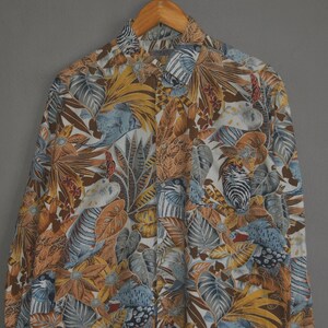 May include: A white button-up shirt with a tropical print featuring a variety of leaves, flowers, and birds in shades of brown, green, blue, and yellow.