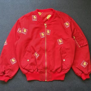 May include: A bright red bomber jacket featuring a gold-toned front zipper, ribbed collar, cuffs, and hem. The exterior is decorated with multiple small square patches, each displaying a yellow chick graphic and the text "THE BEST".