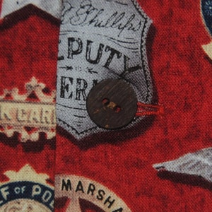 Pode incluir: Close-up de um tecido vermelho com um padr&atilde;o de distintivos de pol&iacute;cia vintage e texto. Um bot&atilde;o grande e redondo de madeira &eacute; costurado no tecido. O tecido tem um aspeto ligeiramente envelhecido.