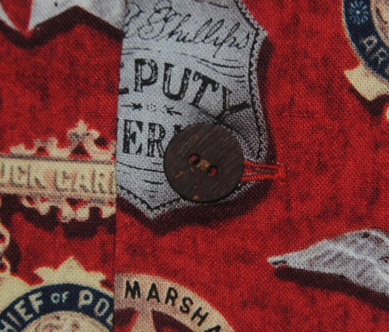 Pode incluir: Close-up de um tecido vermelho com um padr&atilde;o de distintivos de pol&iacute;cia vintage e texto. Um bot&atilde;o grande e redondo de madeira &eacute; costurado no tecido. O tecido tem um aspeto ligeiramente envelhecido.