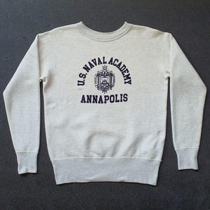 May include: A light gray crewneck sweatshirt with the text "U.S. NAVAL ACADEMY ANNAPOLIS" in navy blue. The design includes a crest. The sweatshirt has long sleeves and a ribbed hem and cuffs.