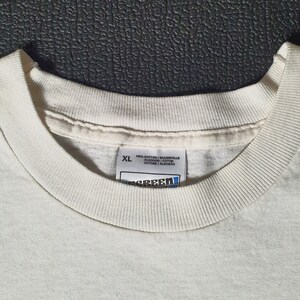 Pode incluir: Uma camiseta branca com uma etiqueta branca que diz "XL 100% Algod&atilde;o / Baumwolle / Algod&oacute;n / Cotone / Algod&atilde;o" e uma etiqueta azul que diz "Screen Star".