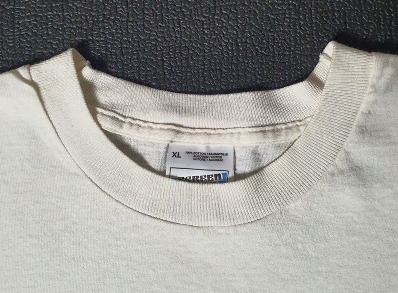 Pode incluir: Uma camiseta branca com uma etiqueta branca que diz "XL 100% Algod&atilde;o / Baumwolle / Algod&oacute;n / Cotone / Algod&atilde;o" e uma etiqueta azul que diz "Screen Star".