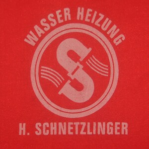 Op de afbeelding: Een rode stoffen patch met witte tekst die "Wasser Heizung" luidt en een witte cirkel met een gestileerde letter "S" erin. Onder de cirkel staat de tekst "H. SCHNETZLINGER".