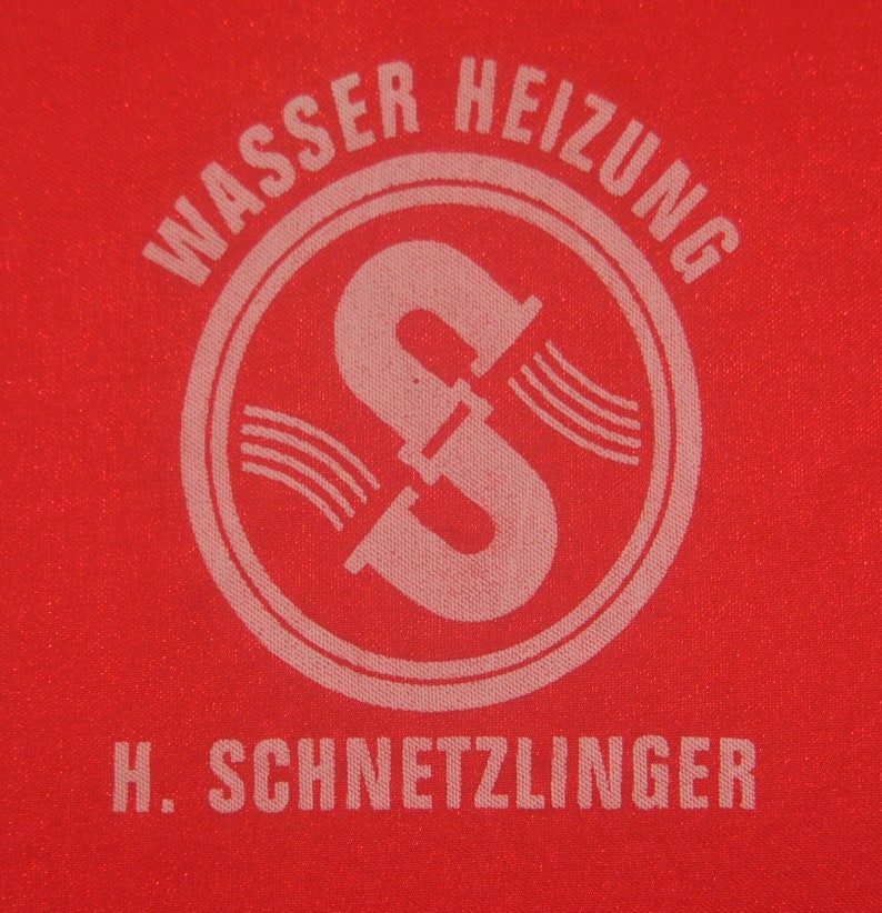 Op de afbeelding: Een rode stoffen patch met witte tekst die "Wasser Heizung" luidt en een witte cirkel met een gestileerde letter "S" erin. Onder de cirkel staat de tekst "H. SCHNETZLINGER".