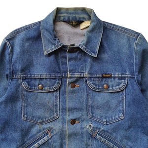May include: Close-up of a vintage blue denim jacket. The jacket features a classic collar, button-down front, two chest pockets with button closures, and the brand name "Wrangler" on a small label. The denim has a faded appearance.