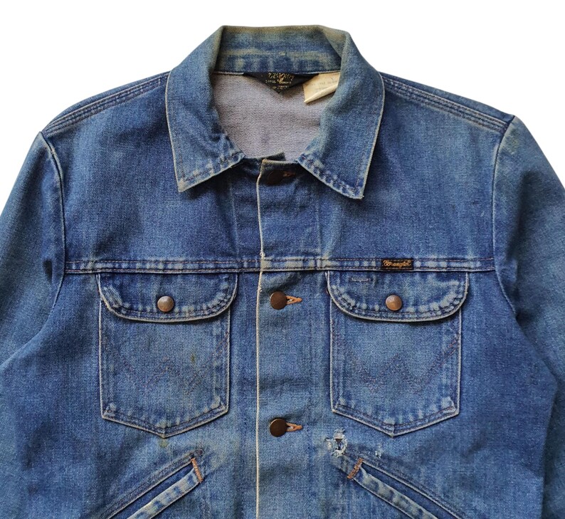 May include: Close-up of a vintage blue denim jacket. The jacket features a classic collar, button-down front, two chest pockets with button closures, and the brand name "Wrangler" on a small label. The denim has a faded appearance.