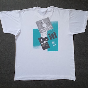 以下が含まれることがあります： 白い半袖Tシャツで、フロッピーディスクのグラフィックプリントが施されています。黒とティール色で「COMPUTER FITNESS... YOU CAN DO IT!」および「AT&T」という文字が印刷されており、ディスクの後ろにはティール色の長方形の背景デザインが含まれています。