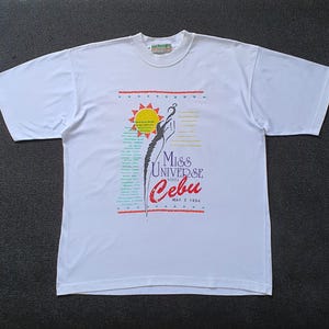 May include: White t-shirt with a colorful graphic design featuring a woman in a swimsuit, a sun, and the text "Miss Universe 1994 Cebu May 21, 1994".
