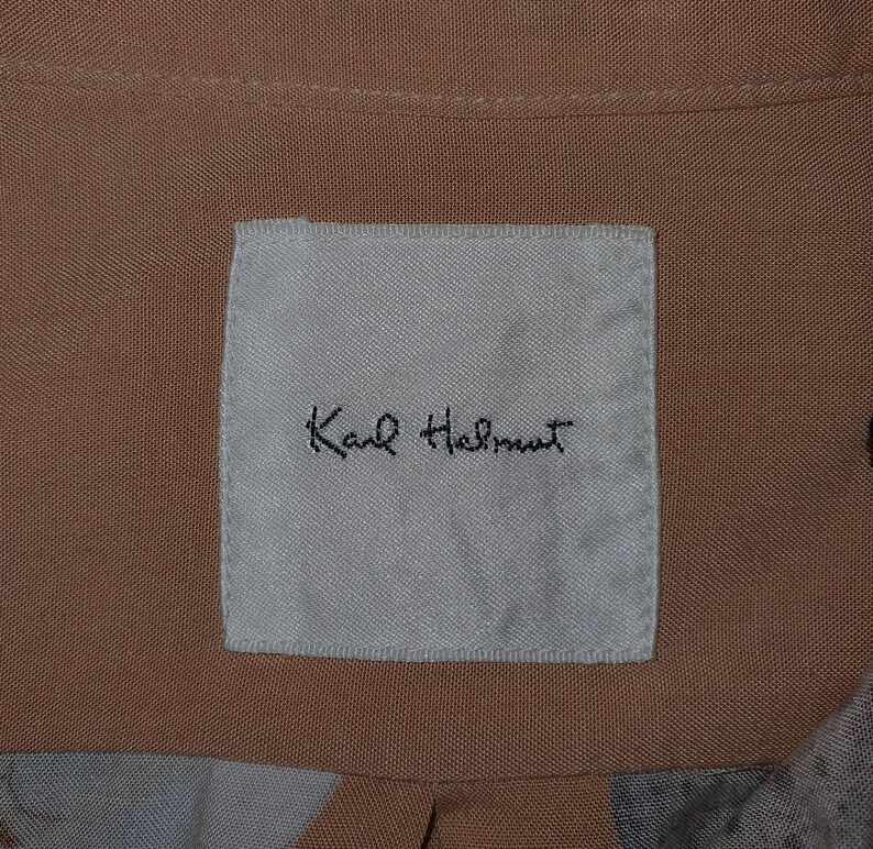 K&ouml;nnte beinhalten: Nahaufnahme eines beigefarbenen Stoffes mit einem wei&szlig;en Etikett. Das Etikett zeigt den handgeschriebenen Text "Karl Helmut" in schwarzer Schrift. Der Stoff hat eine subtile Textur und ist Teil eines Kleidungsst&uuml;cks.