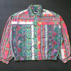 Puede incluir: Chaqueta abotonada colorida con un vibrante patrón geométrico en rojo, verde, morado y blanco. La chaqueta presenta un cuello, mangas largas y una parte delantera abotonada. El nombre de la marca "Jams" es visible en el cuello.