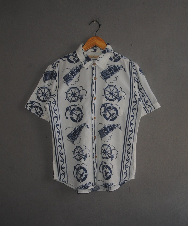 May include: A white short-sleeved button-down shirt with a blue and white nautical print. The print features lighthouses, anchors, and ship wheels.