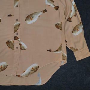K&ouml;nnte beinhalten: Hellbeige Hemd mit Kn&ouml;pfen und einem sich wiederholenden Muster aus Kugelfischen in Braun, Wei&szlig; und Beige. Das lang&auml;rmlige Hemd hat eine einzelne Tasche und eine gekn&ouml;pfte Manschette. Hergestellt aus leichtem Stoff.