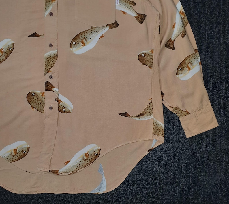 K&ouml;nnte beinhalten: Hellbeige Hemd mit Kn&ouml;pfen und einem sich wiederholenden Muster aus Kugelfischen in Braun, Wei&szlig; und Beige. Das lang&auml;rmlige Hemd hat eine einzelne Tasche und eine gekn&ouml;pfte Manschette. Hergestellt aus leichtem Stoff.