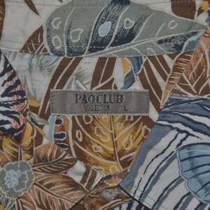 May include: A white button-down shirt with a tropical print featuring brown, blue, and yellow leaves. The shirt has a label that reads "PAOCLUB SELECTION L".