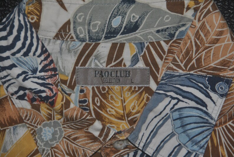 May include: A white button-down shirt with a tropical print featuring brown, blue, and yellow leaves. The shirt has a label that reads "PAOCLUB SELECTION L".