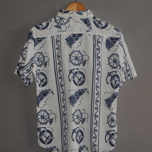 May include: A white short-sleeved button-down shirt with a nautical theme. The shirt features a repeating pattern of blue anchors, ship wheels, and lighthouses.