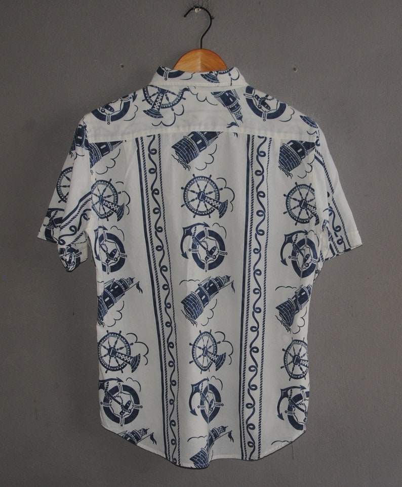 May include: A white short-sleeved button-down shirt with a nautical theme. The shirt features a repeating pattern of blue anchors, ship wheels, and lighthouses.
